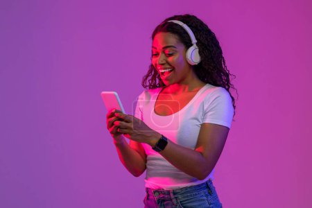 Téléchargez les photos : Excited Black Young Woman Wearing Wireless headphones Using Smartphone In Neon Light, Happy Surprised African American Female Browsing New Application Or Messaging With Friends, Copy Space - en image libre de droit