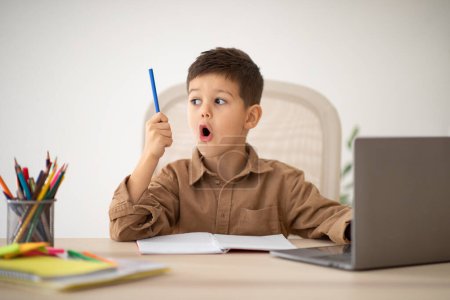 Photo for Happy surprised small european boy sit at table studying, drawing with laptop, looks at pencil with open mouth in room interior. Got idea, creation solution, art lesson and school, learning at home - Royalty Free Image