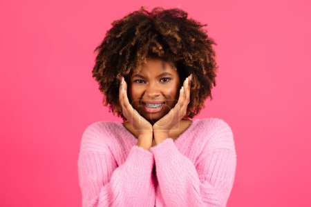Téléchargez les photos : Sad dissatisfied smiling young black curly lady with braces in casual presses hands to cheeks, suffering from toothache, isolated on pink background, studio. Health care, dentistry, caries and pain - en image libre de droit