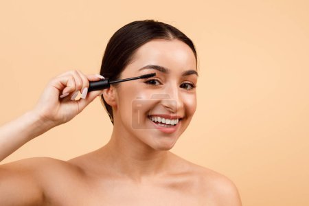 Photo for Portrait Of Beautiful Smiling Young Indian Woman Applying Mascara On Eyelashes, Attractive Hindu Female Making Daily Makeup, Standing With Bare Shoulders Over Beige Studio Background, Copy Space - Royalty Free Image