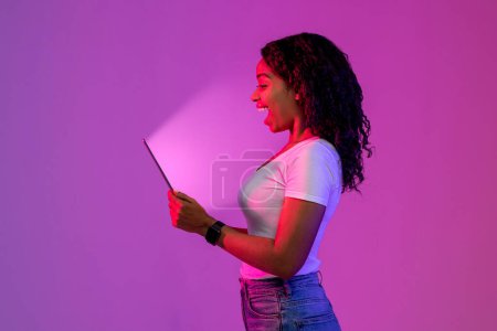 Téléchargez les photos : Surprised Black Woman Looking At Digital Tablet With Glowing Screen, Young African American Lady Playing Online Games Or Browsing Internet On Computer, Standing In Neon Light Over Purple Background - en image libre de droit