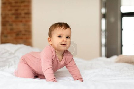 Téléchargez les photos : Adorable baby girl crawling on bed and looking at camera in bedroom interior, free space. Happy childhood concept. Little kids full length portrait - en image libre de droit