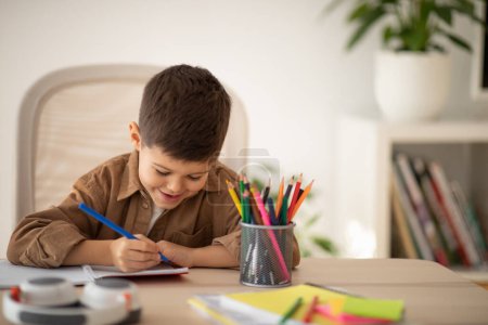 Téléchargez les photos : Busy cheerful cute small european boy sitting at table, draws picture in room, children garden or school interior, free space. Art, study, learning at home and childhood, education due new normal - en image libre de droit