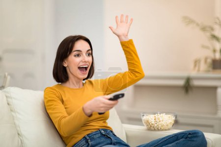 Foto de Emotional happy attractive young woman sitting on couch in cozy living room, watching TV show, football game with popcorn, holding remote controller, gesturing, spending time alone at home, copy space - Imagen libre de derechos