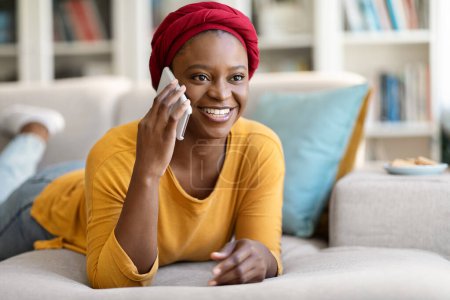 Foto de Happy cheerful young black lady in casual wearing red turban reclining on couch at home, talking on phone with friend or lover, sharing news, looking at copy space and smiling - Imagen libre de derechos