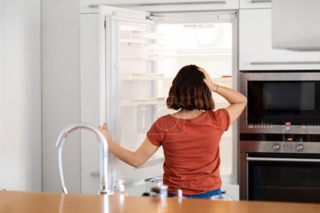 Foto de Hungry Young Woman Looking Inside Of Fridge With Empty Shelves And No Food, Confused Brunette Female Opening Refrigerator And Scratching Head, Starving Lady Suffering Life Crisis, Rear View - Imagen libre de derechos
