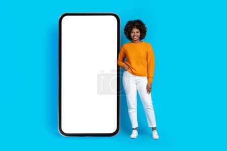 Téléchargez les photos : Cheerful smiling pretty curly millennial african american woman in stylish outfit posing by huge phone with white blank screen, showing nice online offer, blue background, mockup, full length - en image libre de droit