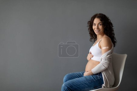 Photo for Portrait Of Smiling Young Pregnant Woman Posing After Coronavirus Vaccination, Expectant Female Sitting On Chair Over Grey Background, Touching Belly And Demonstrating Arm With Adhesive Bandage - Royalty Free Image