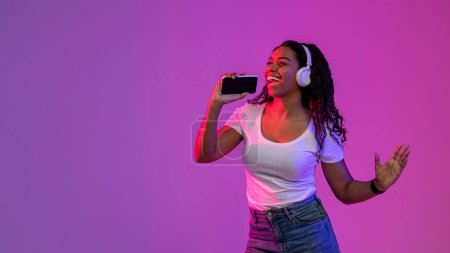Téléchargez les photos : Joyful black woman listening music in wireless headphones and singing with smartphone, cheerful african american female using mobile phone as microphone, standing under neon purple lighting - en image libre de droit
