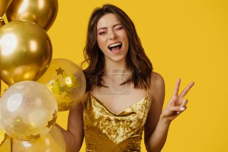 Foto de Funny young lady with birthday balloons winking and gesturing peace, standing on yellow studio background. Playful caucasian woman having party or holiday celebration - Imagen libre de derechos