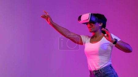 Photo for Virtual Reality Experience. Excited Black Woman Playing Video Game In VR Glasses, Amazed African American Female Standing In Vivid Neon Light Over Purple Studio Background, Panorama With Copy Space - Royalty Free Image