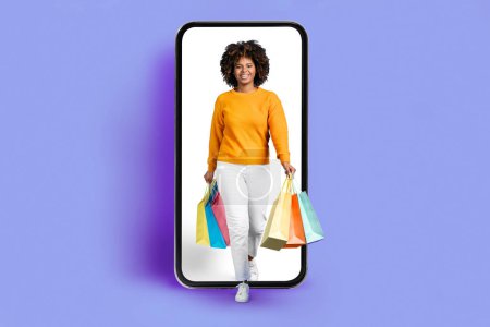 Téléchargez les photos : Pretty cheerful young african american woman in casual with bushy hair shopaholic walking out of big phone with white empty blank screen, smiling, carrying shopping bags, purple background, copy space - en image libre de droit