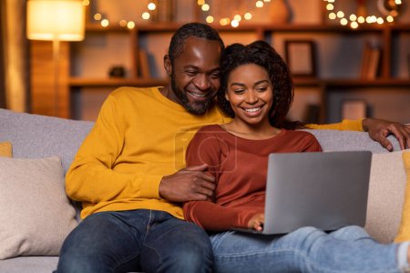 Photo for Happy cheerful loving african american couple sitting on couch, using laptop at cozy winter evening, choosing new furniture, planing vacation, resting at home together, free space - Royalty Free Image