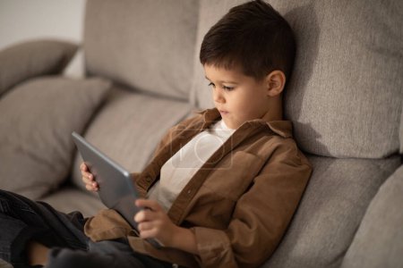 Foto de Serious small european boy sitting on sofa watch video and playing game with tablet in living room interior. Study learning at home with device, childhood and education remotely, entertainment and app - Imagen libre de derechos