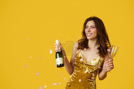 Téléchargez les photos : Festive mood concept. Happy caucasian woman in evening dress with champagne bottle and filled glass in hand posing over yellow studio background with falling confetti - en image libre de droit