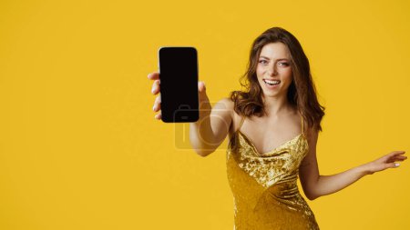 Foto de Happy young caucasian woman wearing shiny golden dress and showing cellphone with blank screen, standing over yellow background, panorama, mockup - Imagen libre de derechos