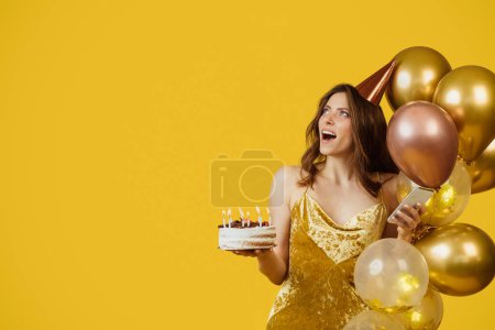 Foto de Surprised woman with balloons, yummy cake and smartphone, looking aside at free space on yellow studio background. Pretty lady celebrating anniversary, feeling happy and excited - Imagen libre de derechos