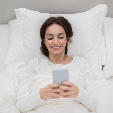 Photo for Top view of cheerful happy beautiful young brunette woman wearing white pajamas using cell phone in bed after waking up in the morning, chatting with boyfriend, bedroom interior, copy space - Royalty Free Image