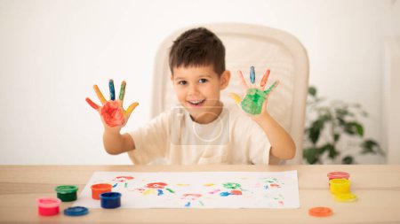 Téléchargez les photos : Cheerful caucasian little child sits at table draws with finger paints, painted his palms in room interior. Fun alone, entertainment and play at home, art, fantasy and childhood, study, education - en image libre de droit