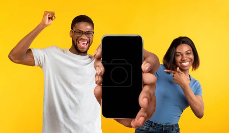 Téléchargez les photos : Excited Black Man And Woman Demonstrating Big Smartphone With Blank Screen With Copy Space For App Or Website Design, Cheerful Couple With Cellphone Standing On Yellow Background, Collage, Mockup - en image libre de droit