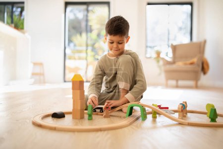 Foto de Cheerful small european boy in pajamas sits on floor plays with road and cars, enjoy spare time in nursery room interior. Entertainment, fun alone, education, childhood at home, lifestyle and creative - Imagen libre de derechos