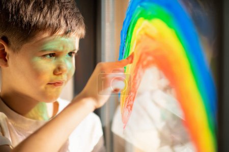 Téléchargez les photos : Smiling cute european small kid draws rainbow on window with finger and paint, close up, sun flare. Fun alone, fantasy at home and kindergarten, childhood and art, education and creative, lifestyle - en image libre de droit