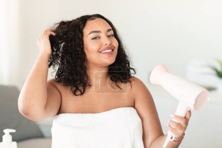 Foto de Beautiful oversize woman in bath towel using hairdryer after morning shower and smiling at camera, copy space. Body positive lady drying her hair, making hairdo. Everyday hygiene concept - Imagen libre de derechos