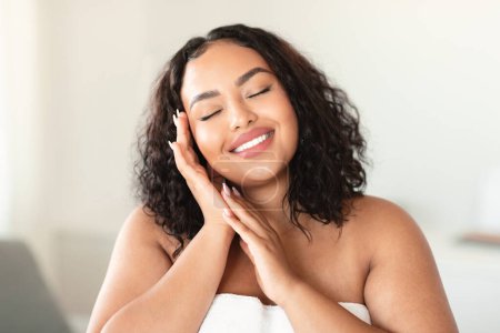 Foto de Beautiful happy overweight lady touching her face and smiling with closed eyes, positive bodypositive woman standing wrapped in towel in bathroom interior - Imagen libre de derechos
