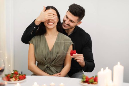Téléchargez les photos : Engagement, anniversary celebration, love, relationships concept. Loving man surprising his girlfriend with valentine present red gift box, happy young couple celebrating lovers day at home - en image libre de droit