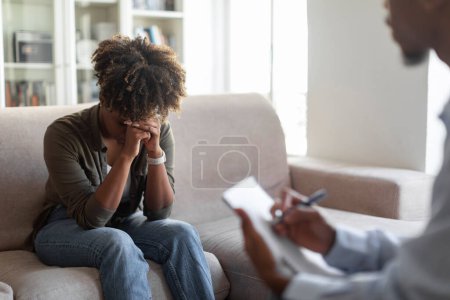 Photo for Upset young black woman in casual sitting on couch, cover face with palms, crying while have therapy session with man therapist, african lady fighting addiction, visiting psychologist, copy space - Royalty Free Image