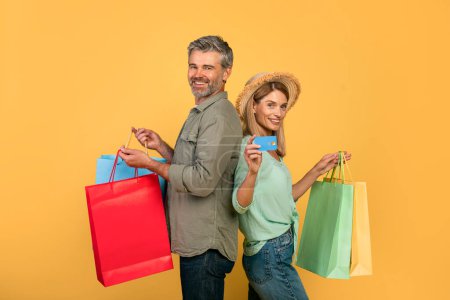 Téléchargez les photos : Portrait of middle aged caucasian spouses showing credit card and holding colorful shopper bags after purchases in shopping mall, standing over yellow background, studio shot - en image libre de droit