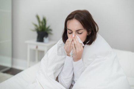 Foto de Sixk unhappy millennial brunette lady in pajamas sitting in bed covered in warm blanket at home, sneezing, using napkin, suffering from cold, flu or coronavirus concept, copy space, closeup - Imagen libre de derechos