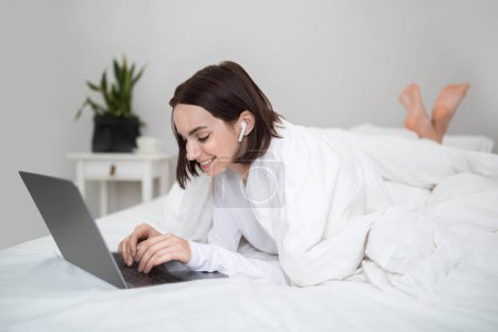 Téléchargez les photos : Happy cheerful joyful young brunette woman freelancer working from home, lying on bed under cover, typing on laptop keyboard, using wireless earpods, copy space. Freelance, remote job concept - en image libre de droit