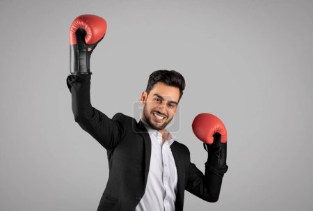 Téléchargez les photos : Handsome Arab Businessman Wearing Boxing Glowes And Celebrating Success, Overjoyed Middle Eastern Male Entrepreneur In Suit Emotionally Reacting To Win, Posing Over Grey Studio Background, Copy Space - en image libre de droit