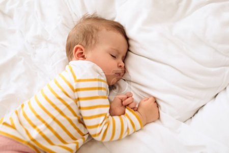 Photo for Cute little baby girl sleeping on side, lying in bed on white bedding, above view, free space. Peaceful child napping, resting during daytime sleep - Royalty Free Image