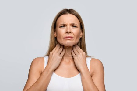 Foto de Sad unhappy adult middle aged blonde woman touching her neck with both hands, lady suffering from sore throat, have pain while swallowing, isolated on grey studio background, copy space - Imagen libre de derechos