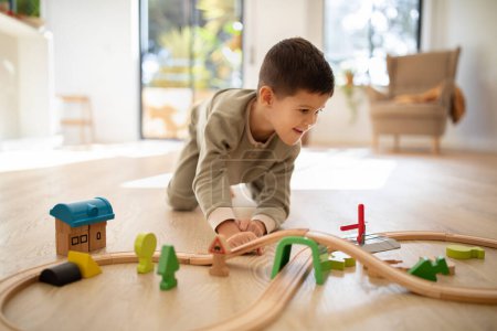 Foto de Glad cheerful small european boy in pajamas on floor plays with road and cars alone, enjoy fantasy at spare time in children room interior, sun flare. Childhood, entertainment with toys, fun at home - Imagen libre de derechos