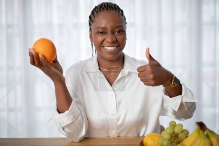 Téléchargez les photos : Positive cheerful good-looking attractive young black lady in white shirt showing fresh juicy orange and thumb up at camera, sitting at desk next to window. Healthy lifestyle, diet, nutrition concept - en image libre de droit