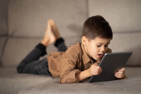 Foto de Concentrated european small kid uses tablet to watch video, plays educational games, enjoy spare time, lies on sofa in living room interior, empty space. App for fun, video call at home, addiction - Imagen libre de derechos