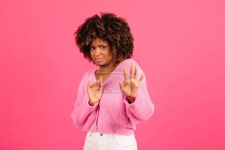 Téléchargez les photos : Sad frightened young black curly lady in casual fencing off with hands, making stop sign, isolated on pink background, studio, copy space. Fear, horror, gesture from domestic violence, discrimination - en image libre de droit