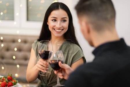 Foto de Attractive brunette long-haired hispanic young woman with light makeup in nice dress have date, cheering glasses of red wine with her boyfriend, festive background decorated with lights, copy space - Imagen libre de derechos