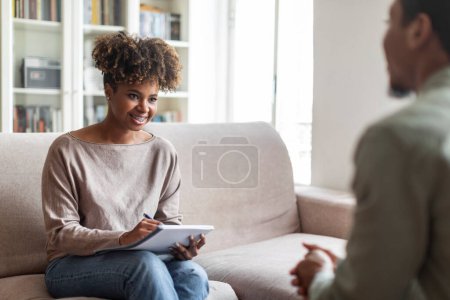 Foto de Mental health. Friendly cheerful attractive youg african american woman therapist have conversation with male patient, taking notes in notepad and smiling, counselor office interior, copy space - Imagen libre de derechos