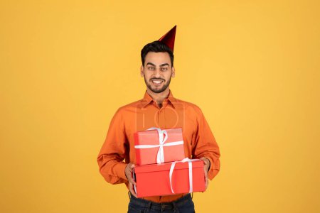 Téléchargez les photos : Handsome arab man holding wrapped gift boxes and wearing birthday hat while standing on yellow studio background, happy middle eastern male having B-day party, carrying presents, copy space - en image libre de droit