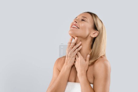 Foto de Happy smiling attractive half-naked middle aged blonde woman covered in towel touching her neck, enjoying anti-aging SPA salon treatment results, grey studio background, copy space - Imagen libre de derechos