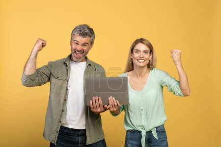Photo for Overjoyed middle aged couple celebrating win, holding laptop, screaming and gesturing over yellow studio background. Caucasian man and woman gambling and trading online - Royalty Free Image