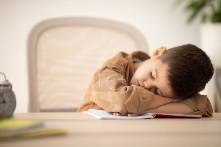 Tired small european boy lies at table sleeping, resting from lesson in school room interior. Fatigue, overwork, boredom from art, learning at home and children garden, childhood and upbringing