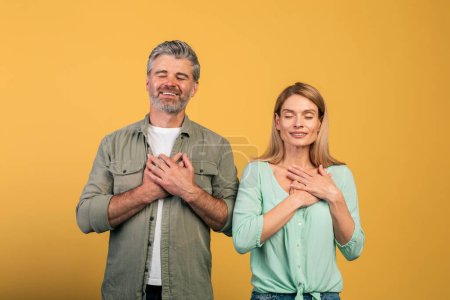 Téléchargez les photos : Thank you. Grateful middle aged couple with closed eyes keeping both hands on chest, expressing grattitude and kindness, standing over yellow background, studio shot - en image libre de droit