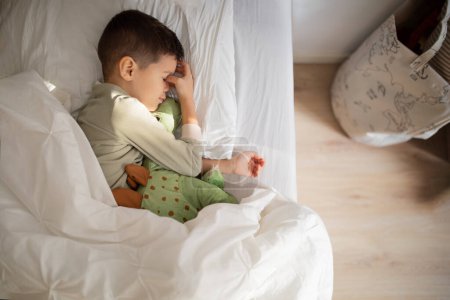 Foto de Tired european small kid in pajamas sleeps, wakes up on bed with dinosaur toy in bedroom interior. Sweet sleep and dreams, good morning, comfort for child, rest and relaxation, childhood at home - Imagen libre de derechos