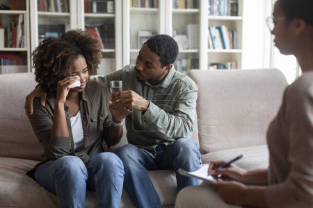 Foto de Loving attentive young black husband hugging comforting his crying spouse wife girlfriend suffering from depression while having family therapy with woman psychologist, guy giving glass of water - Imagen libre de derechos