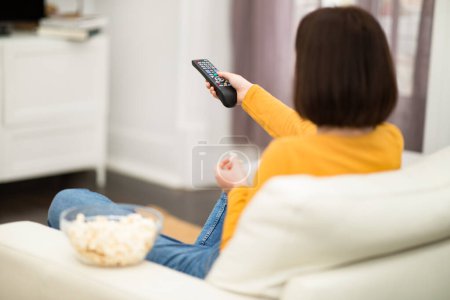 Foto de Unrecognizable brunette young woman in comfy casual outwear sitting on couch in living room, holding remote, eating popcorn, switching channels on TV, spending weekend at home, back view, copy space - Imagen libre de derechos
